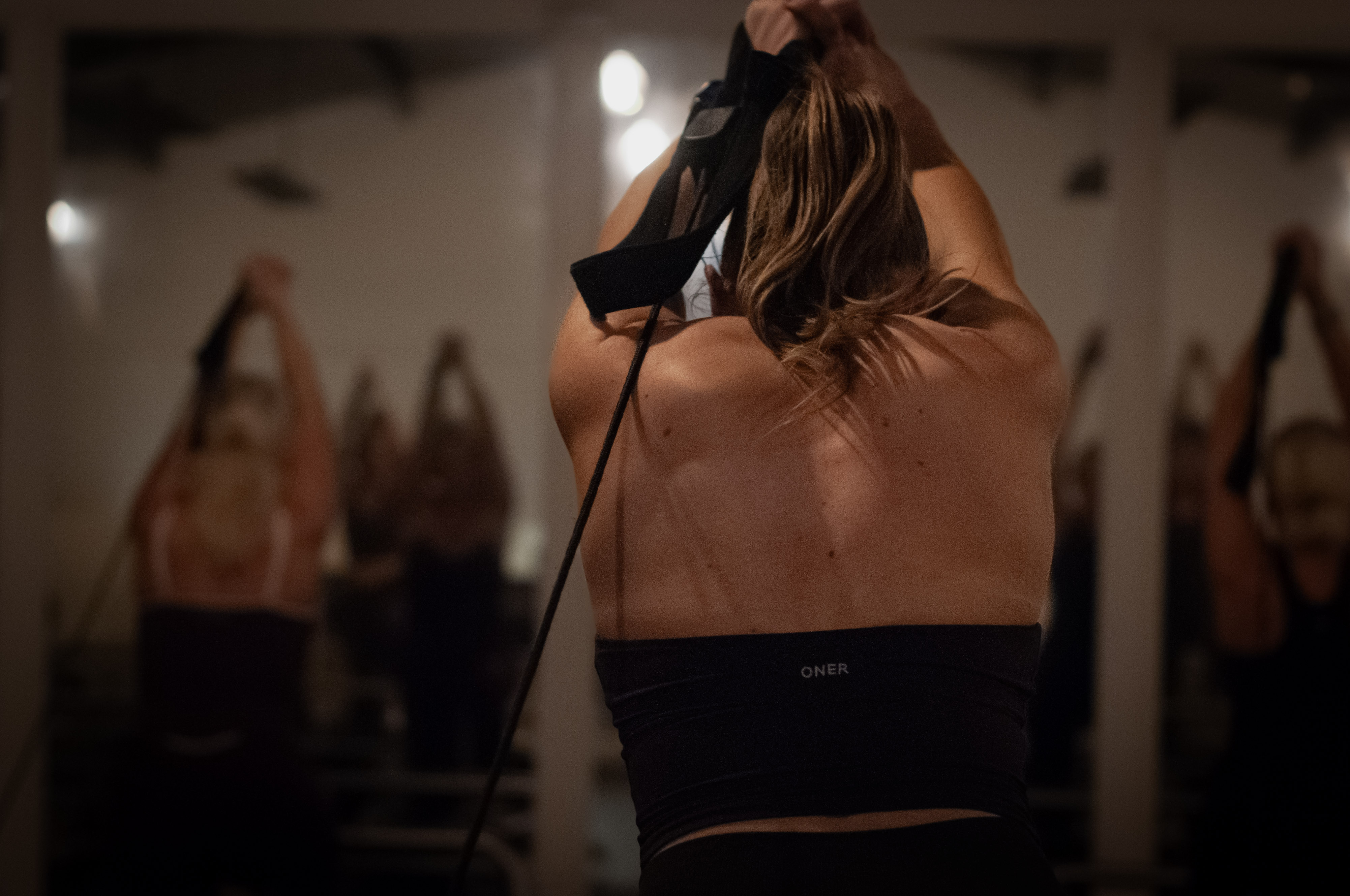 A student stretching with her arms overhead, with other students behind her in the warm yoga room