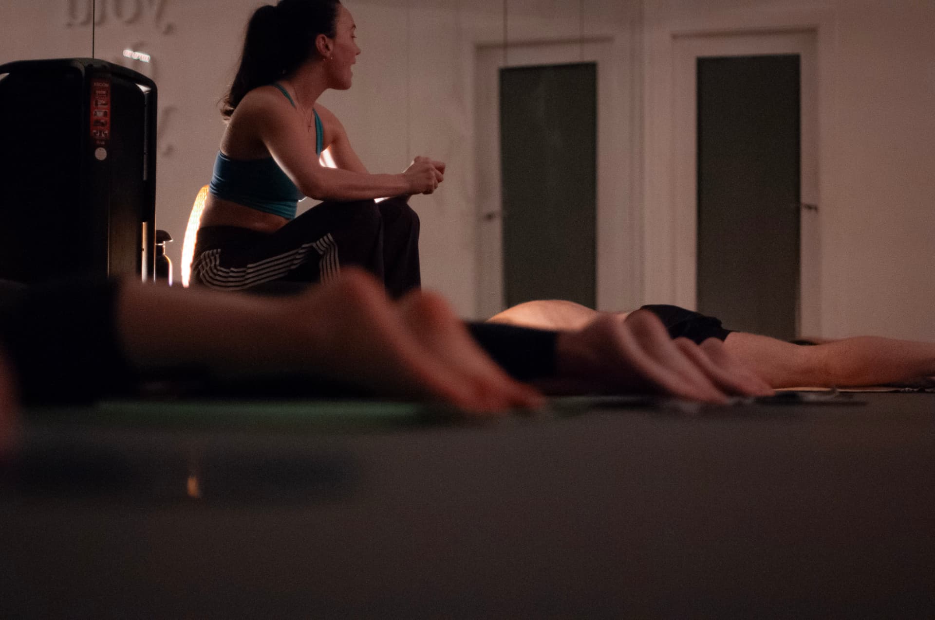 The instructor seated in the warm, low-lit room as the class rests along their mats